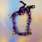 Full view of a wide purple and white patterned bandana bracelet, adorned with three charms (an evil eye, a heart, and stars) and secured with a black lobster clasp and chain