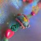 Extreme close-up of the Chunky Bead Charm in the center, featuring light blue clay and small pink and yellow hearts, next to a green smiley face bead