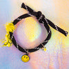 Full view of a black rope bracelet with white accents, tied with a knot, featuring a yellow smiley face charm, a small gold star charm, and a decorative yellow fabric tie