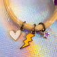 Close-up of the charms on the beige cord bracelet, clearly showing the white enamel heart, the bright yellow lightning bolt, and the purple enamel star charm