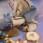 Macro view focusing on a gold-rimmed white enamel charm with a small gold heart and a tan tassel, attached to the frayed denim fabric of the necklace