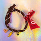 Full view of a ebony rope bracelet with white accents, tied with a knot, featuring three gold-toned charms: a peach heart, a yellow lightning bolt, a black star, and a decorative red bandana fabric tie