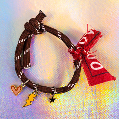 Full view of a ebony rope bracelet with white accents, tied with a knot, featuring three gold-toned charms: a peach heart, a yellow lightning bolt, a black star, and a decorative red bandana fabric tie