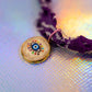 Detailed shot of the round, gold-rimmed charm featuring a colorful, modern evil eye design set in white enamel, attached to the textured purple fabric bracelet
