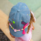 side angle of denim cap showing neon heart pin and bead band