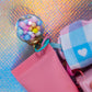 Macro shot of the Gumball Charm (a clear sphere filled with pastel pink, blue, and yellow mini balls) next to the pink lip balm prop and blue/pink gingham ribbon