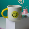 White ceramic mug with yellow handle and smiling face print on front