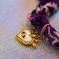 Macro shot of the beautiful gold-toned heart charm with a white enamel center and pink/clear rhinestones, dangling from the purple patterned bandana bracelet