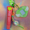 Full view of the neon green winged heart charm, peach sorbet lip butter balm, and colorful Kandi style bead string, embodying Cyber Y2K aesthetics