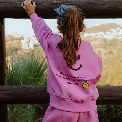 Girl wearing bubble gum pink kids fleece jogger set with smiley graphic on back, standing by wooden fence outdoors