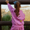 Girl wearing bubble gum pink kids fleece jogger set with smiley graphic on back, standing by wooden fence outdoors