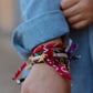 Close-up on a person's wrist wearing multiple cord bracelets (red, green, brown, beige) with charm accents and red bandana fabric ties