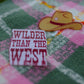 Close-up of cowboy hat pin and “Wilder Than The West” slogan pin on cotton candy and galaxy green plaid scarf