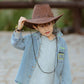 Kid adjusting cowboy hat while wearing Pocket Full of Dreams denim shirt
