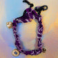 Full product shot showing the purple fabric bracelet with black chain closure and all three charms, displayed against a shimmery, iridescent background