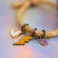 Detailed shot focusing on the three enamel charms, white heart, yellow lightning bolt, and purple star attached to the sand-colored cord bracelet