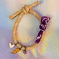 Full view of a sand-colored rope bracelet with white accents, tied in a knot, featuring three gold-toned charms (white heart, yellow lightning bolt, purple star) and a purple patterned fabric tie
