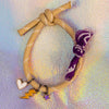 Full view of a sand-colored rope bracelet with white accents, tied in a knot, featuring three gold-toned charms (white heart, yellow lightning bolt, purple star) and a purple patterned fabric tie