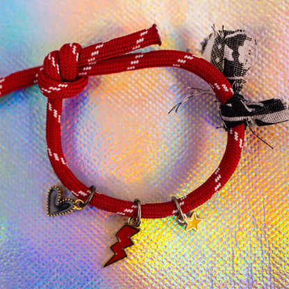 Full view of a bright red rope bracelet with white accents, tied in a knot, featuring three gold-toned charms (black heart, red lightning bolt, yellow star) and a decorative black/white fabric tie