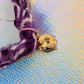 Close-up of the tiny gold-toned round charm featuring three sparkling stars, attached to the knotted purple fabric bandana bracelet.