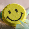 Close-up of yellow smiley face bath bomb with textured fizz surface and bold black facial design for cheerful bath time fun