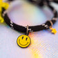 Close-up of the black and white cord bracelet, focusing on the vibrant yellow enamel smiley face charm dangling from a small metal ring